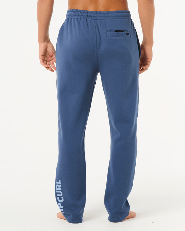 Back view of man wearing blue Rip Curl sweatpants with elastic waistband, back pocket, and logo print on lower leg.