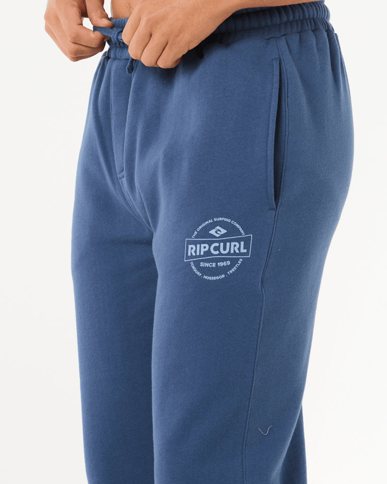 Close-up of blue Rip Curl sweatpants with elastic waistband, drawstring, side pockets, and printed logo on upper leg.