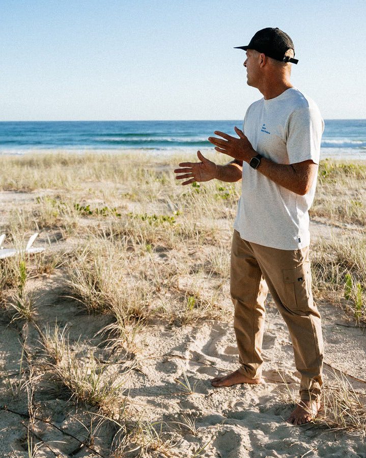 Man wearing tan Billabong pants on sand, styled with a white tee and cap.