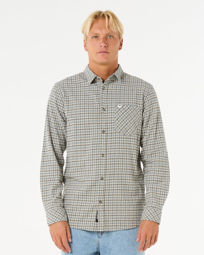 Man wearing a long-sleeve button-up plaid shirt in neutral tones with a front chest pocket, paired with light blue jeans.