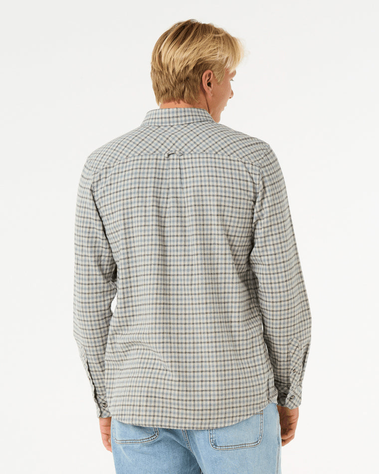 Back view of a man wearing a long-sleeve plaid button-up shirt in neutral tones, paired with light blue denim jeans.