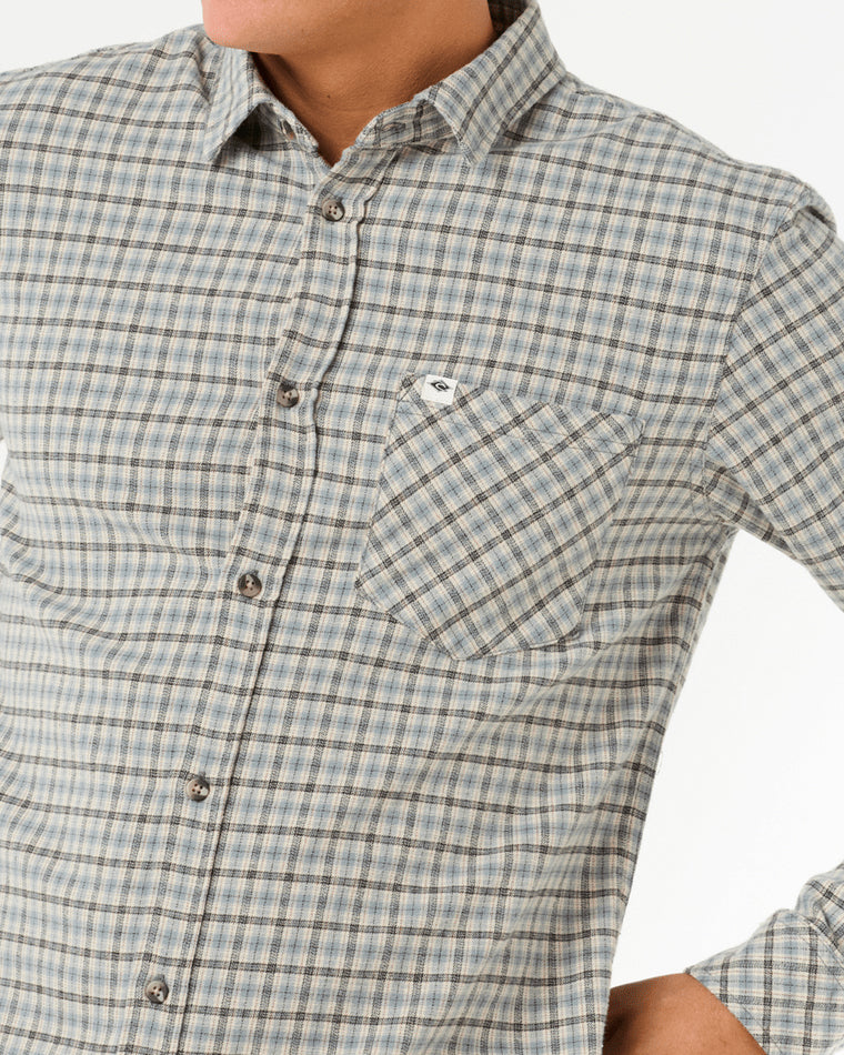 Close-up of a plaid button-up shirt with a chest pocket and small logo tag, featuring a neutral color palette and button details.