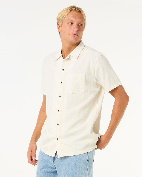 Cream short-sleeve button-up shirt with single chest pocket, styled with light wash denim jeans.