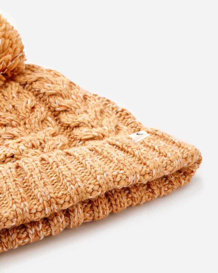 Close-up of a caramel-colored knit beanie with a cable knit pattern, ribbed fold-over cuff, and a small brand tag detail.