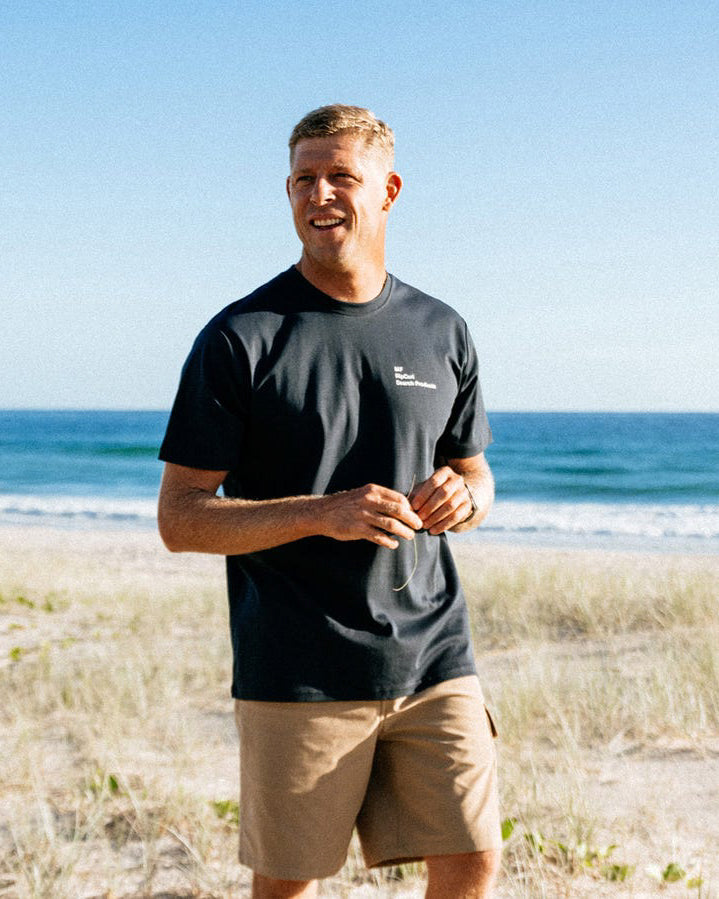 Man wearing navy Rip Curl tee and tan shorts on sand near the ocean.