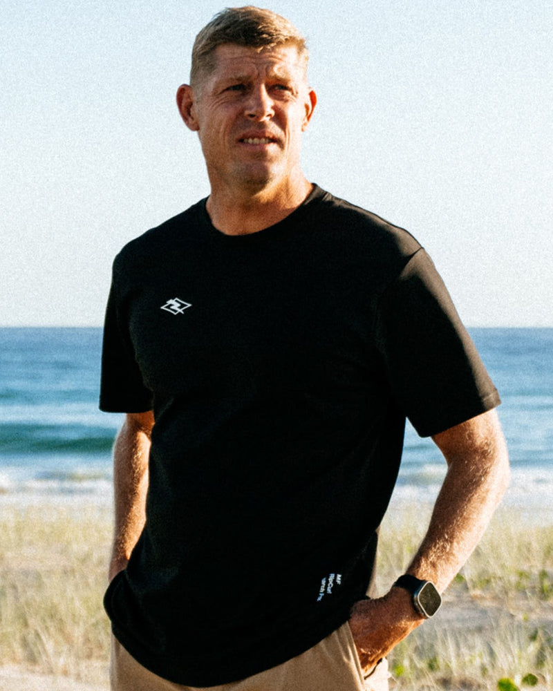 Black short sleeve tee with chest and hem branding, worn casually with beige shorts at the beach.