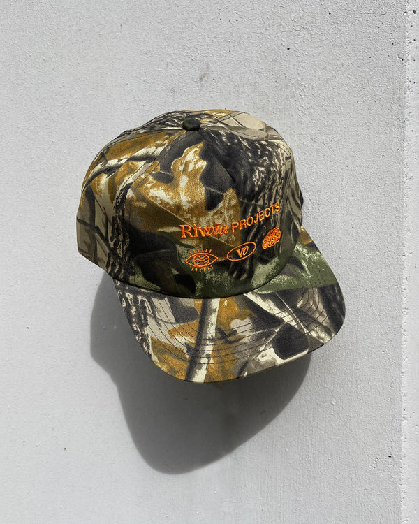 Side view of camo Rivvia Projects cap with orange logo detail