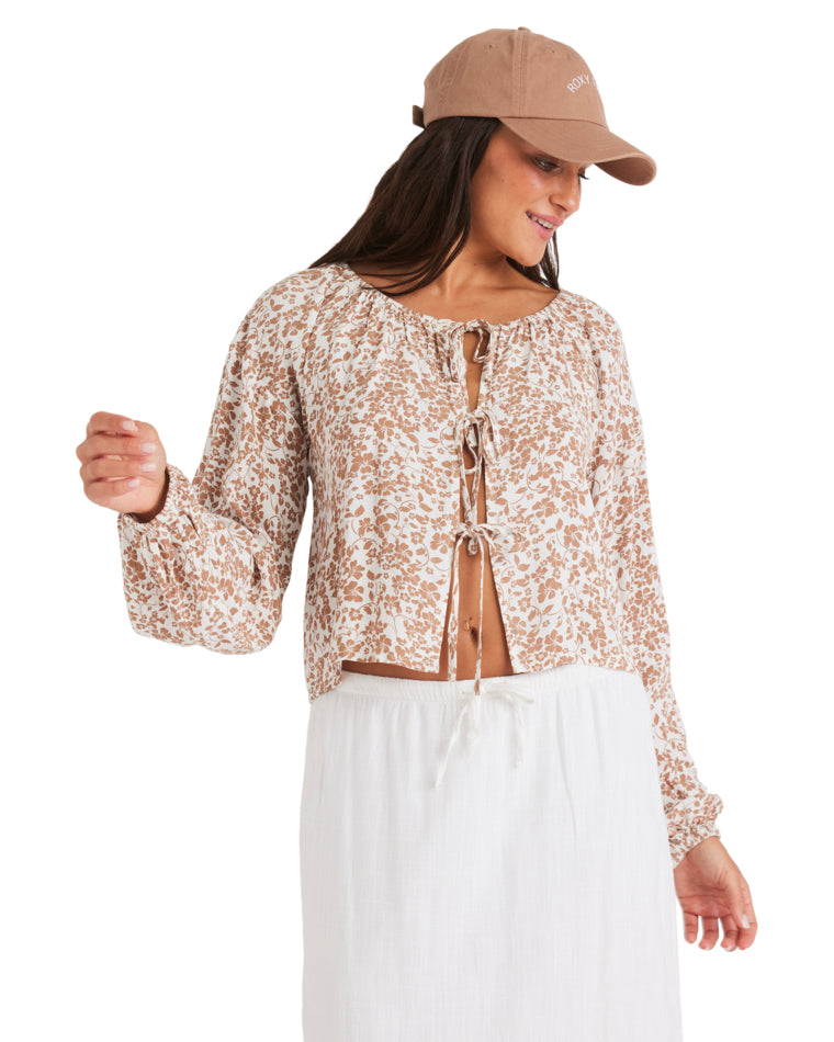 Woman in a light brown printed blouse with front ties and white skirt, smiling and looking down.