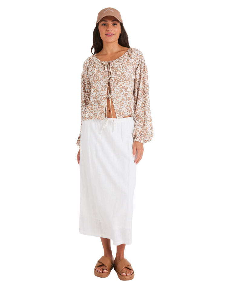 Full-length image of woman in tie-front printed blouse, white drawstring skirt, brown sandals, and a tan cap.