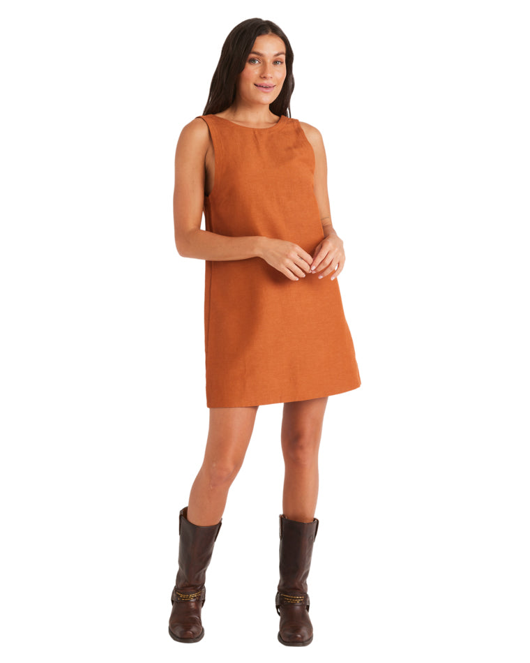 Woman in sleeveless rust mini dress and dark brown boots, standing with hands clasped.