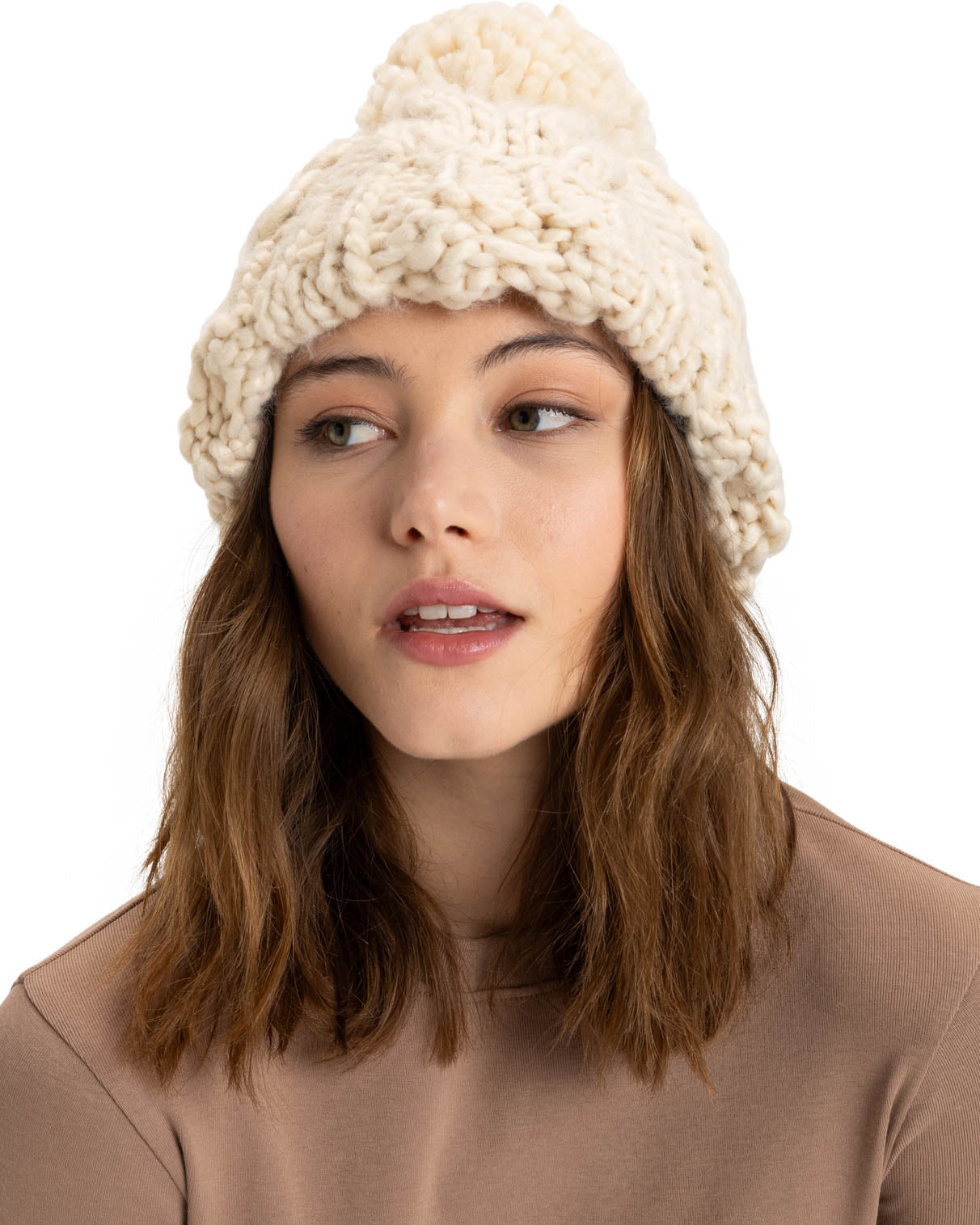 Woman wearing a chunky knit cream beanie with a pom-pom, styled with a neutral brown top, looking slightly to the side.
