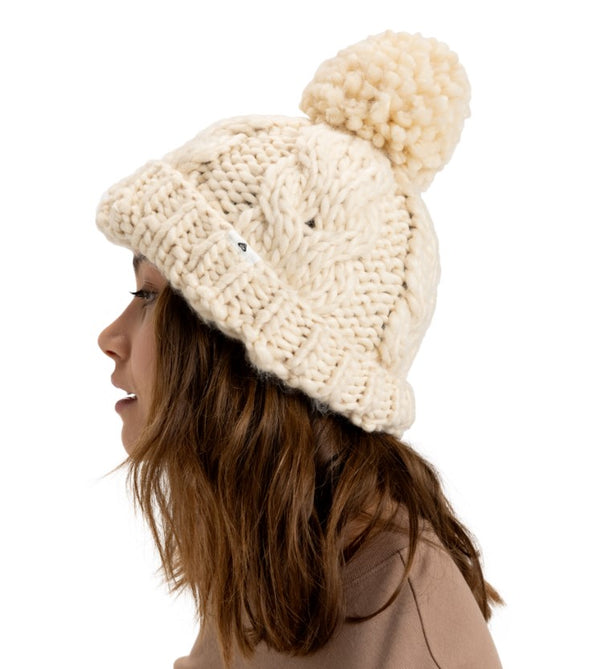 Side profile of a woman wearing a chunky knit cream beanie with a pom-pom, styled with a neutral brown top.