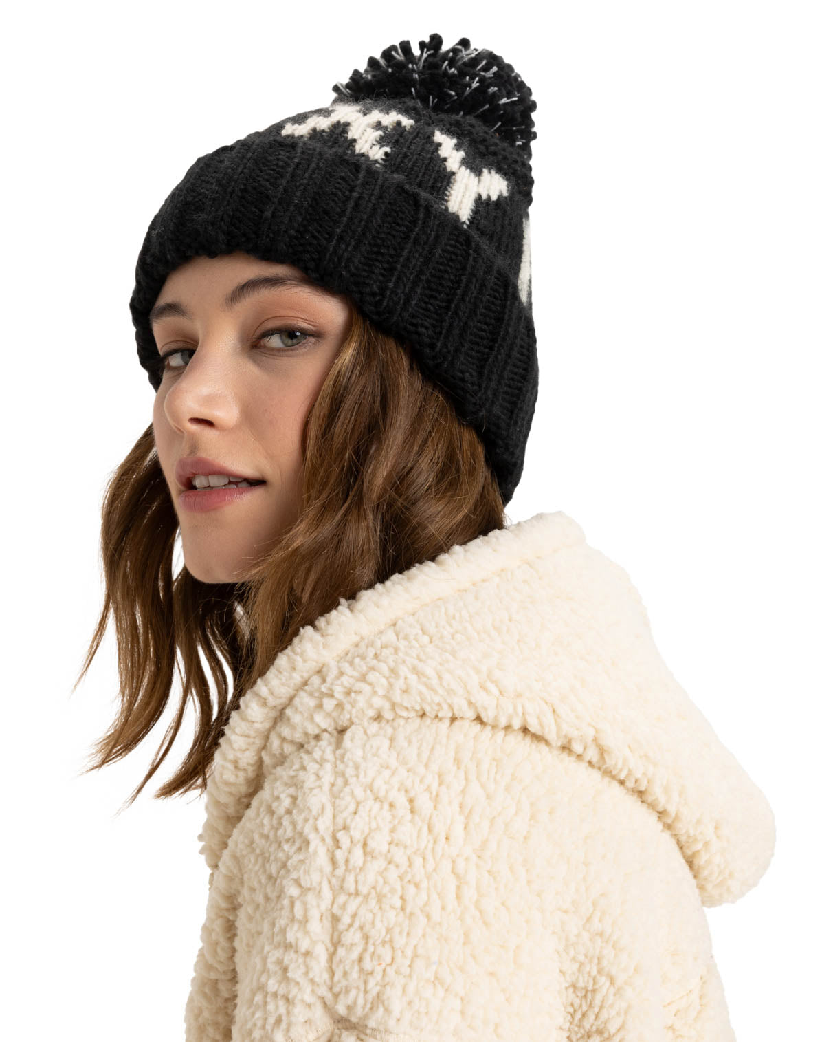 Black knitted beanie with white text and a pom-pom on top, worn by a model in a cream sherpa jacket, side profile view.