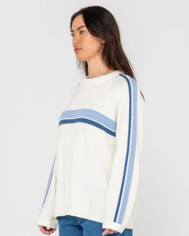 Side view of a woman wearing a cream knit sweater with blue stripe details across the chest and down the sleeves