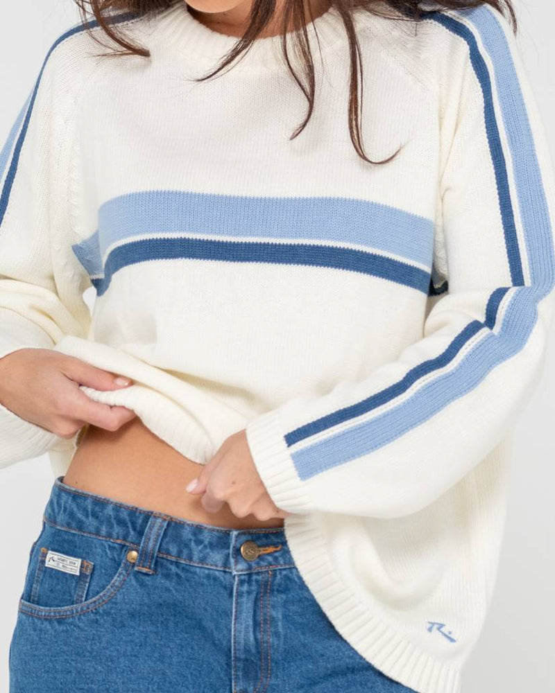 Close-up of a woman wearing a cream knit sweater with blue stripes, slightly lifting the hem to reveal her waistline.