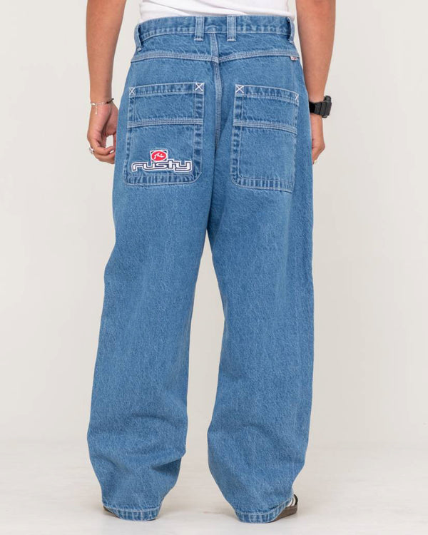 Back view of loose-fit blue denim jeans featuring large patch pockets with Rusty logo embroidery, styled with a white t-shirt.