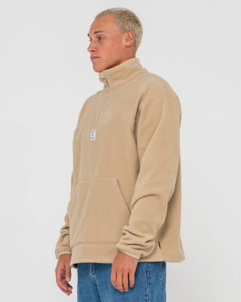 Side view of a man wearing a beige fleece pullover with a half-zip collar and kangaroo pocket, paired with blue jeans.