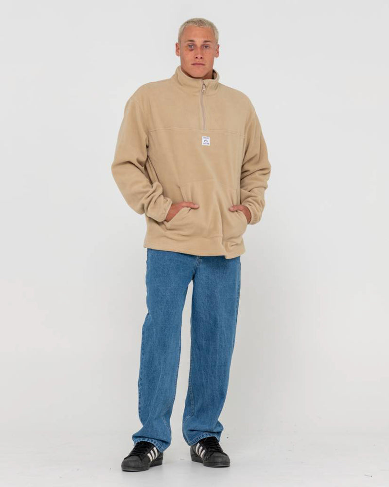 Full-body shot of a man wearing a beige fleece pullover with a half-zip collar, blue jeans, and black sneakers.