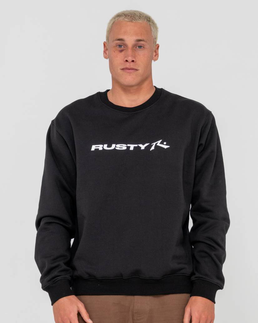Man wearing a black Rusty crewneck sweatshirt with a white logo on the chest, paired with brown pants.