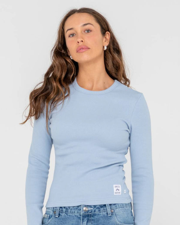 Woman wearing a light blue long-sleeve ribbed top with a crew neckline and a small white logo patch near the hem.