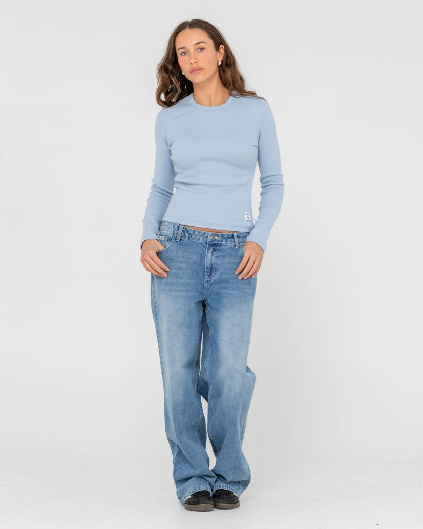 Woman wearing a light blue long-sleeve ribbed top and relaxed-fit blue jeans, standing against a plain white background.