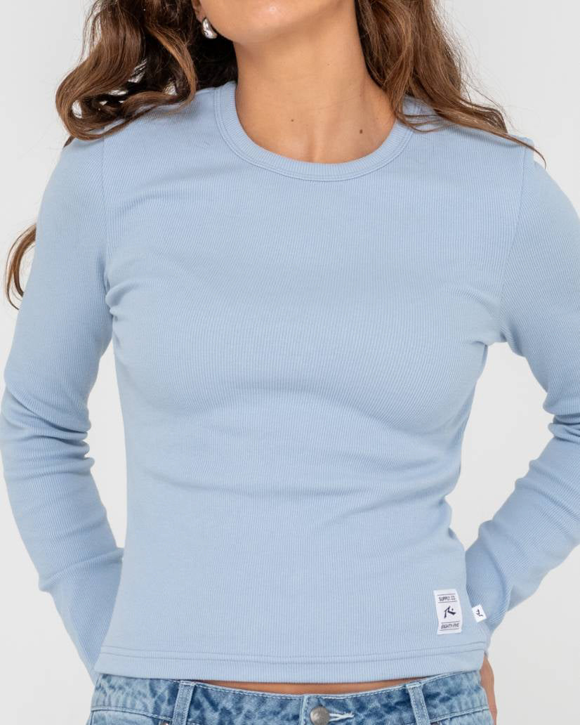 Close-up of a woman wearing a light blue ribbed long-sleeve top with a small logo patch near the hem.