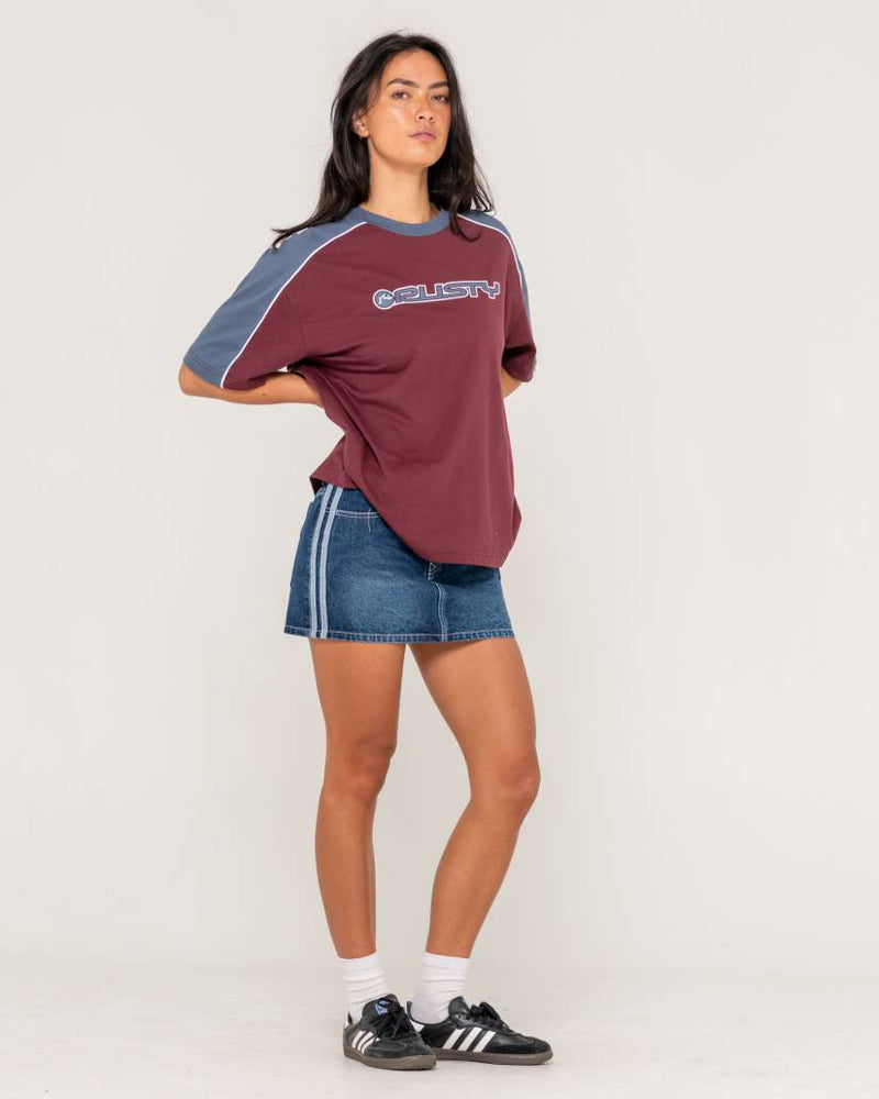 Model wearing a Rusty oversized burgundy T-shirt with navy contrast sleeves, a denim mini skirt, and black Adidas sneakers.