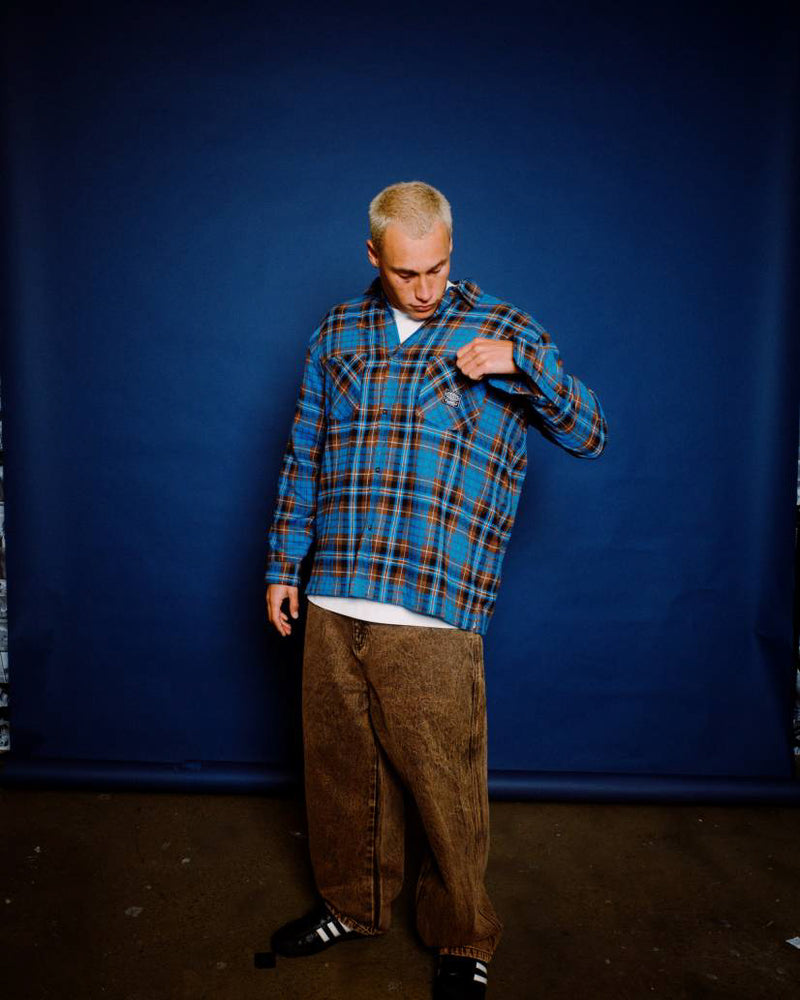 Man wearing a blue and brown plaid flannel shirt over a white tee, paired with brown jeans and black sneakers, posing indoors.