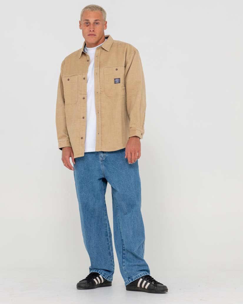 Full-body view of a man wearing a beige corduroy button-up jacket, white t-shirt, blue jeans, and black Adidas sneakers.