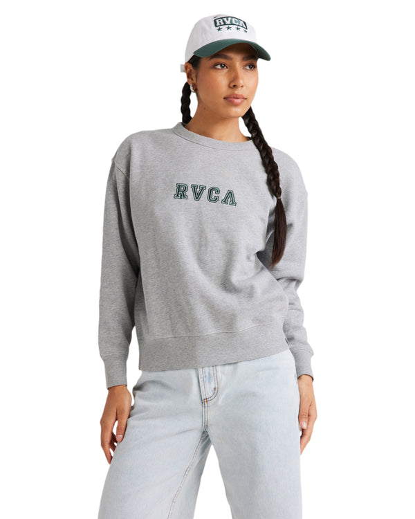 Woman wearing a grey RVCA fleece crew with green embroidered logo, paired with light-wash jeans and a white RVCA cap.