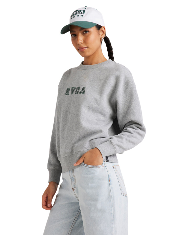 Side view of a woman wearing a grey RVCA fleece crew with green embroidered logo, paired with light-wash jeans and a white cap.