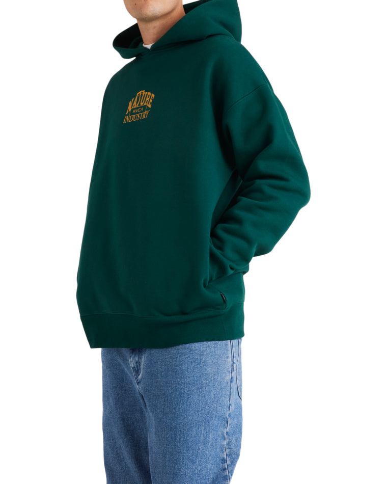 Side view of a person wearing a dark green RVCA hoodie with gold 'Nature Industry' text on the chest, paired with jeans.