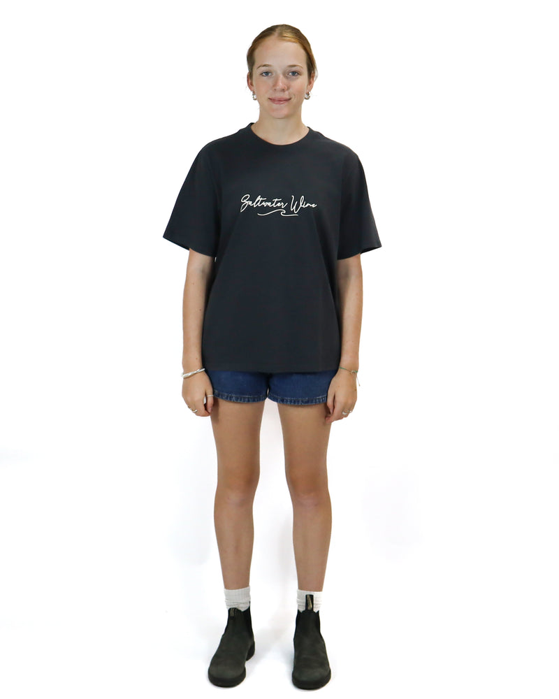 Full-body view of a model wearing a black oversized t-shirt with "Saltwater Wine" text, denim shorts, and black boots.