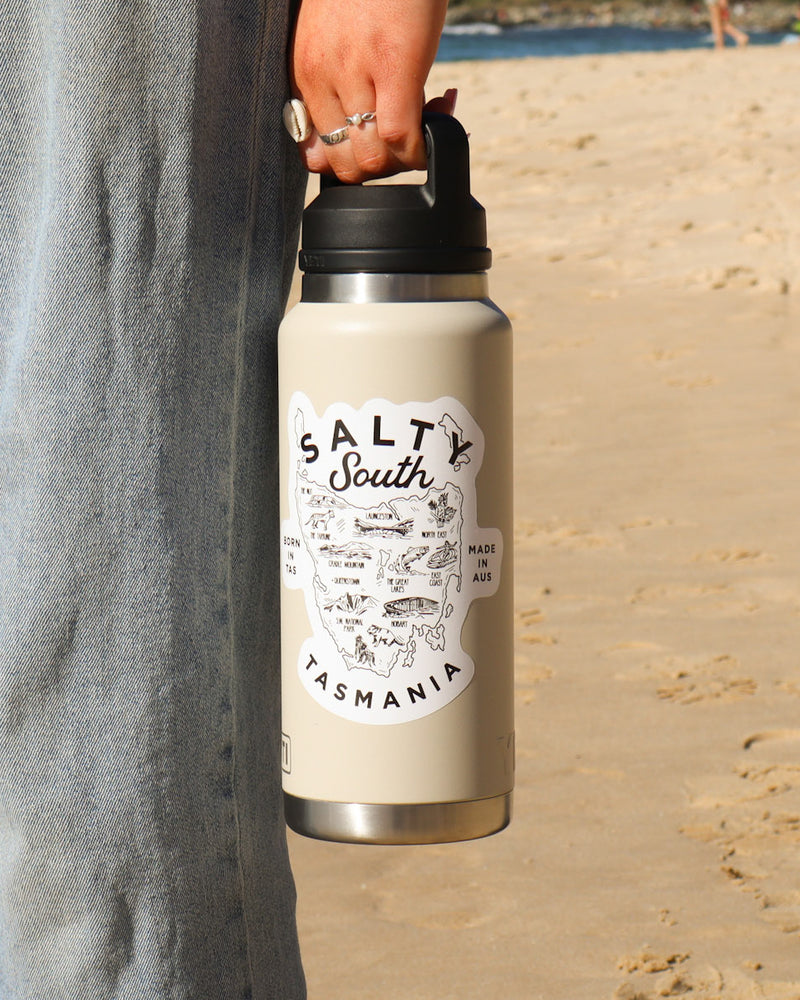 Cream stainless steel drink bottle with Salty South Tasmania map logo, made in Australia and shown held at the beach
