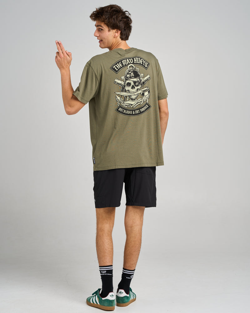 A model wearing an olive green graphic t-shirt with a skull design on the back, paired with black shorts and sneakers.