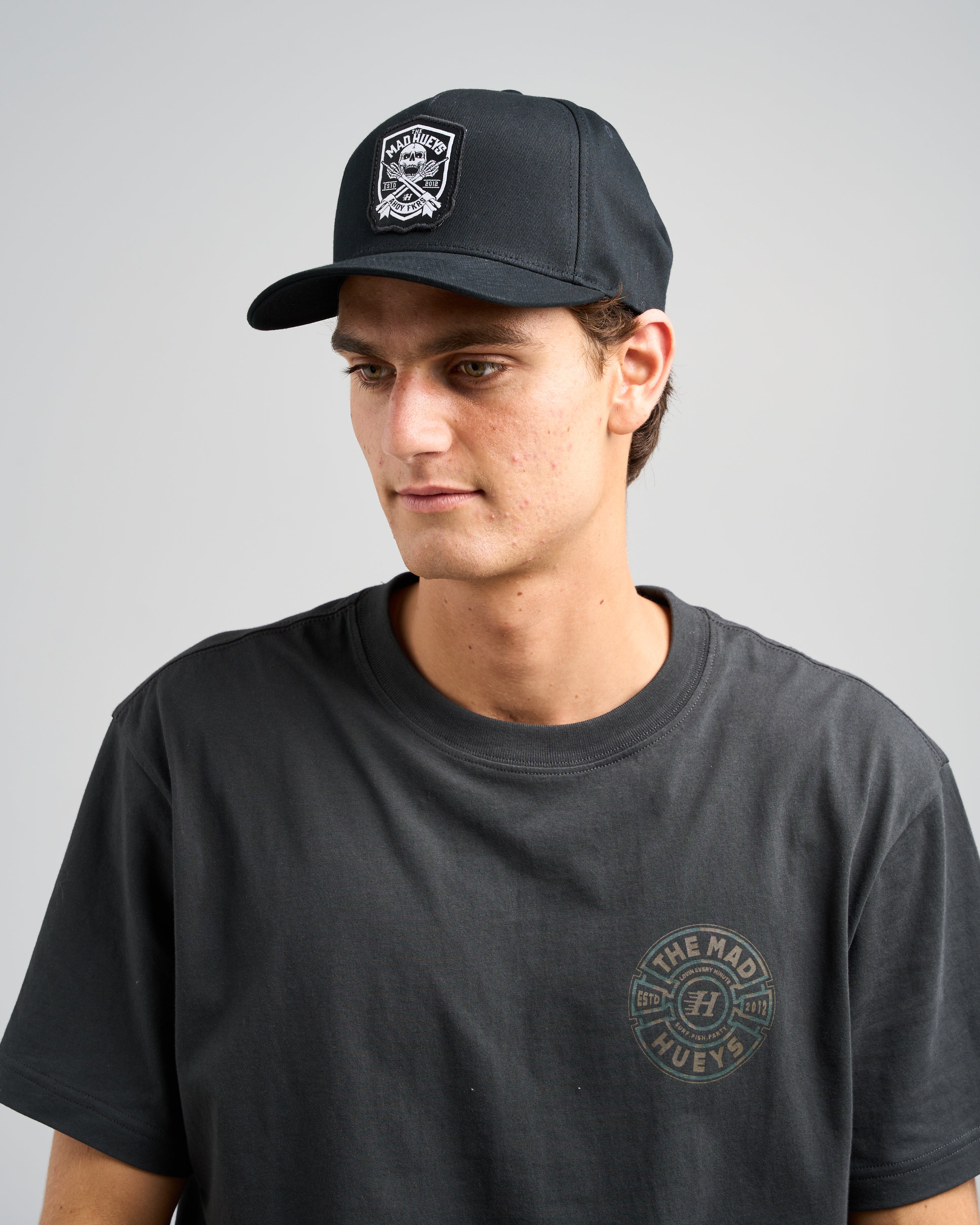 A model wearing a black cap with the "Mad Hueys" logo patch, paired with a black t-shirt featuring subtle graphic text.