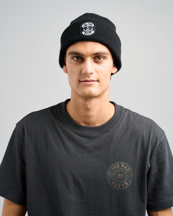 Man wearing a black ribbed beanie with The Mad Hueys logo patch and a dark grey t-shirt featuring a circular chest logo.