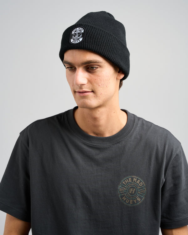 Man wearing a black ribbed beanie with The Mad Hueys logo patch and a dark grey t-shirt featuring a circular chest logo.