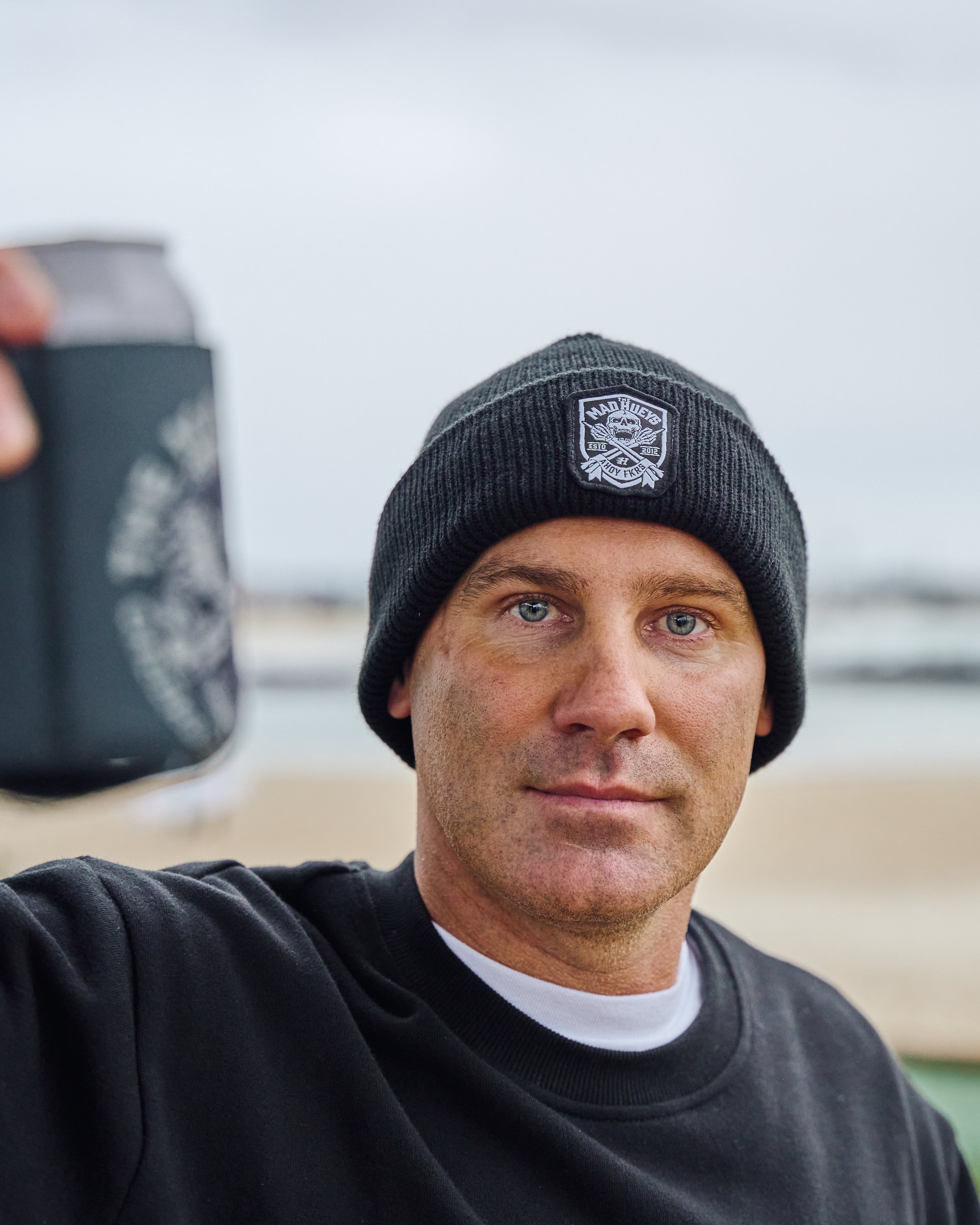 Man wearing a black ribbed beanie with The Mad Hueys logo and a black sweatshirt, holding a drink with a branded cooler.