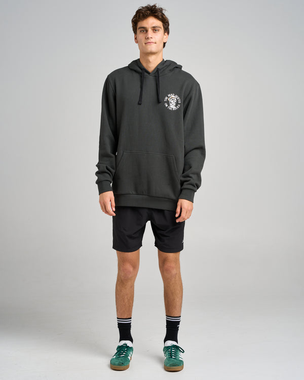 Male model wearing a black hoodie with a small chest logo, black shorts, striped socks, and green sneakers, standing casually.