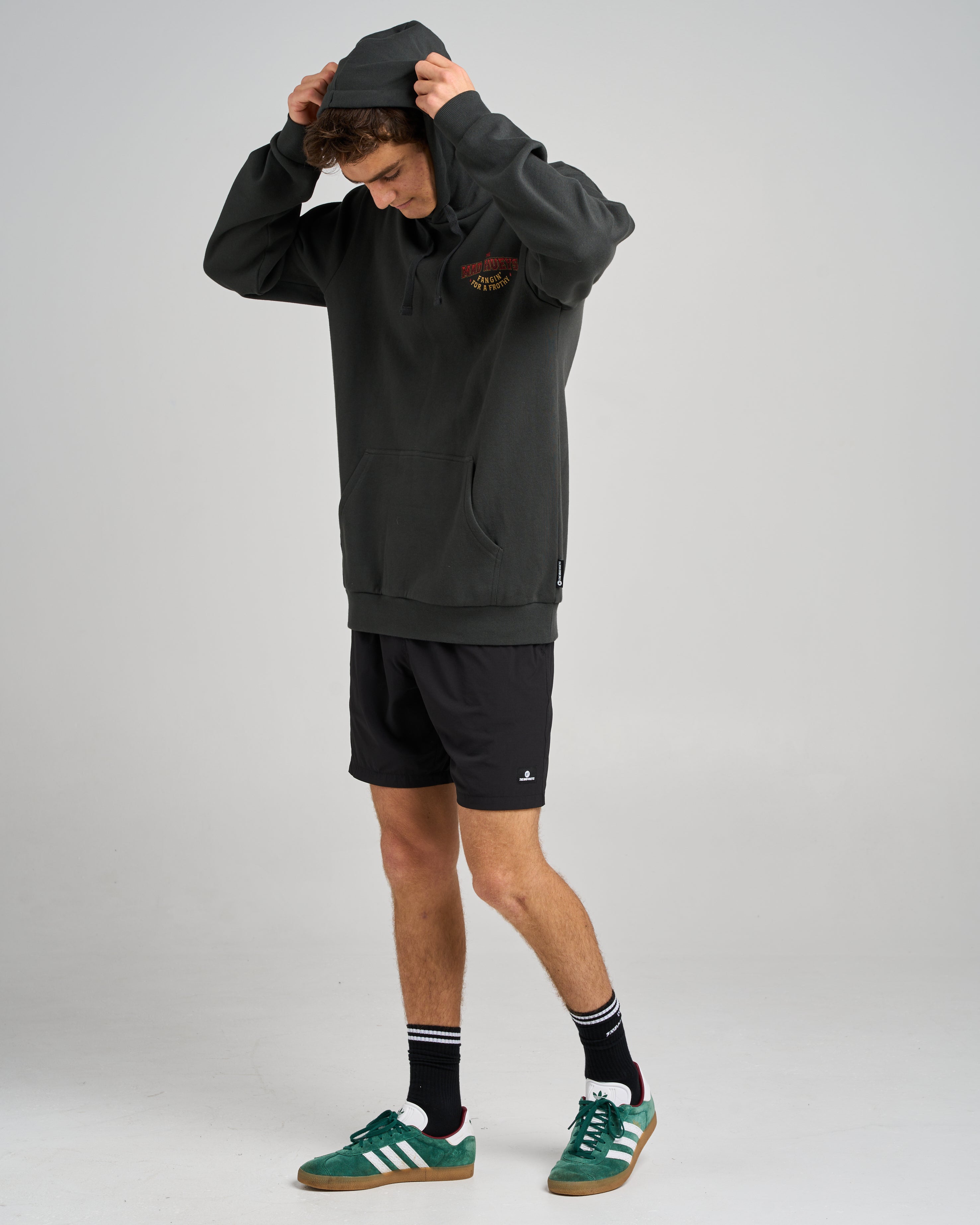 Man wearing black hoodie with small 'The Mad Hueys' chest logo, black shorts, and green Adidas sneakers, adjusting hood.