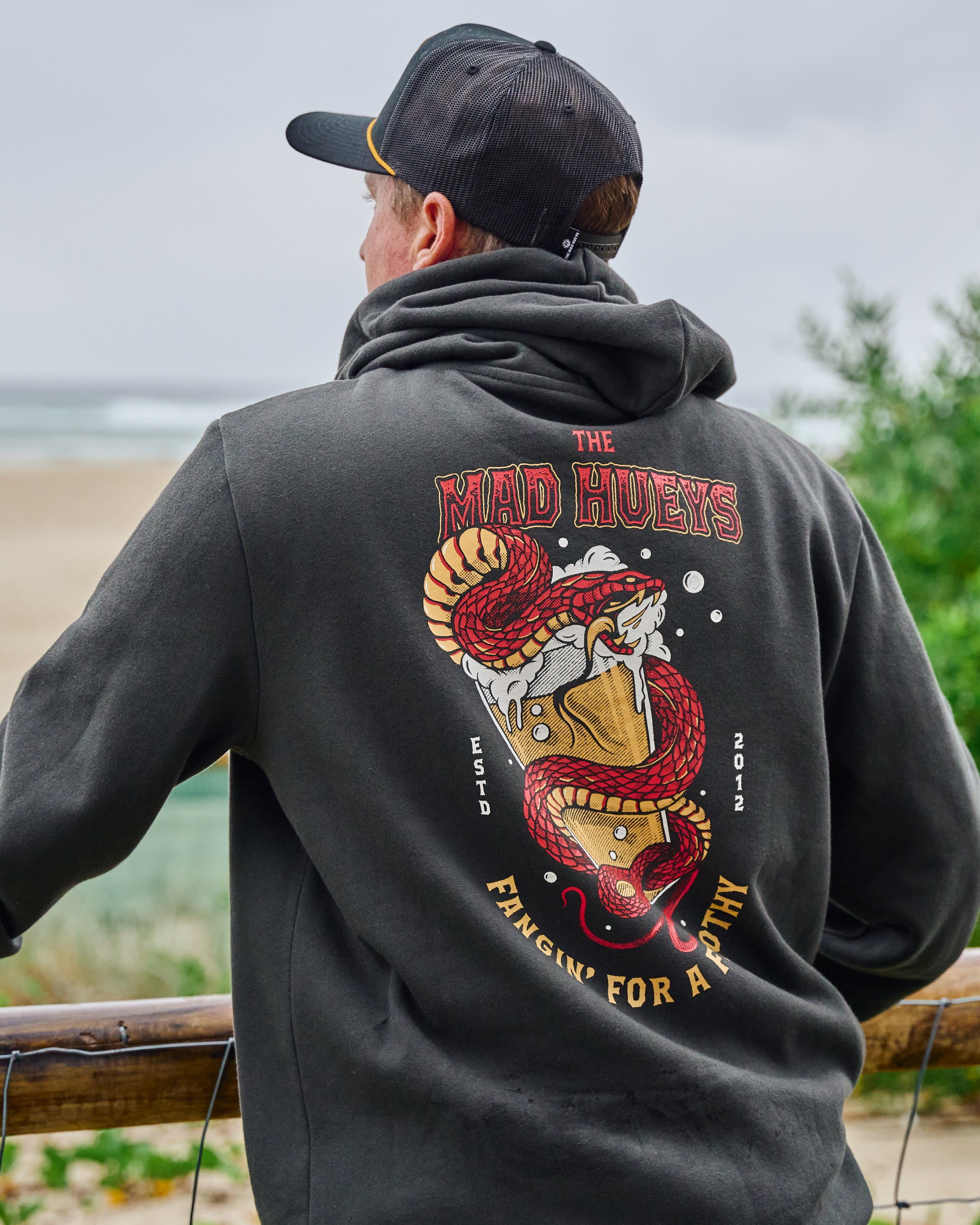 Man wearing black hoodie with 'The Mad Hueys' snake and beer graphic on the back, standing outdoors near a beach railing.