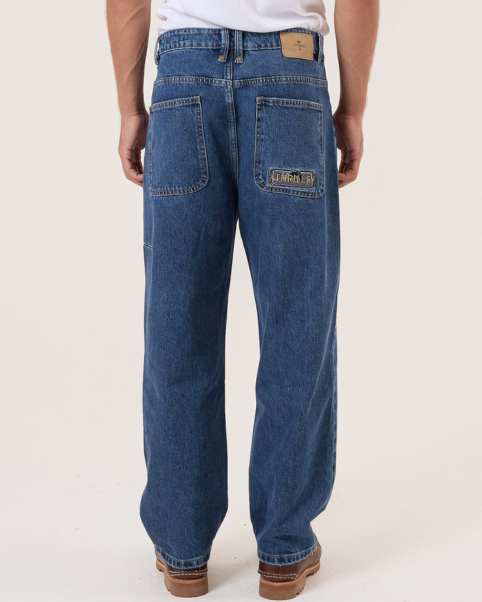 Back view of men's straight-leg blue denim jeans with two back pockets, a branded patch, and brown leather shoes.