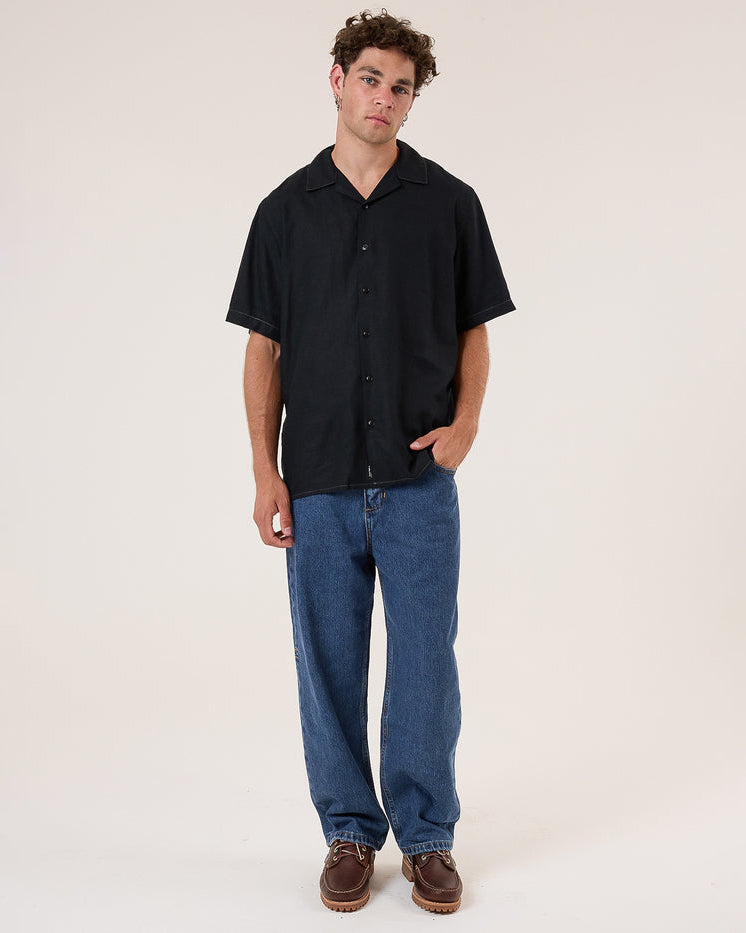 Male model wearing a black short-sleeve button-up shirt, relaxed-fit blue jeans, and brown leather shoes, standing casually.