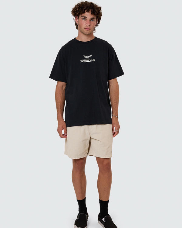 Full body view of model wearing black Thrills t-shirt with beige shorts and black slip-on shoes