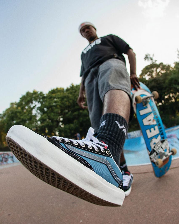 Skater in black Vans shirt and grey shorts holding a blue skateboard, wearing blue and black Vans sneakers at a skatepark.