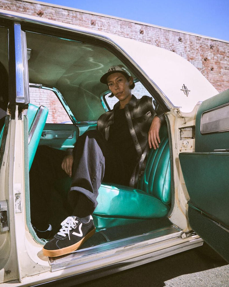 A person wearing black low-top sneakers with white accents, a wavy stripe design, and gum rubber soles, sitting in a vintage car.