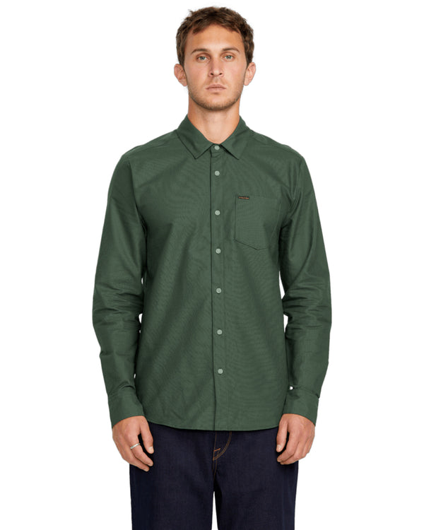 Male model wearing a long-sleeve dark green button-up shirt with a front pocket, paired with dark jeans, standing straight.