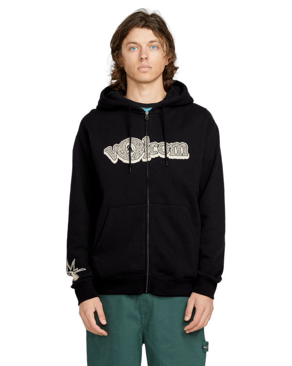 Model wearing a black Volcom zip-up hoodie with a bold logo on the chest, front pockets, and a subtle sleeve graphic detail.