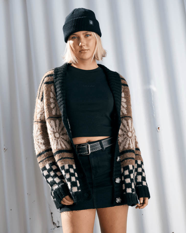 Model wearing an oversized knit cardigan with floral and checkerboard patterns, paired with a black crop top, skirt, and beanie.